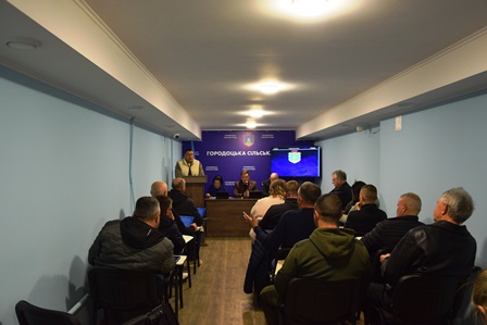 У залі засідань відбувається сесія сільської ради: попереду за столом сидять представники керівництва громади, один із них виступає з доповіддю.