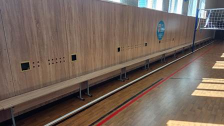 Дерев’яні лавки вздовж стіни у спортивному залі після оновлення. На підлозі видно розмітку для спортивних ігор та волейбольну сітку, освітлення від вікон падає на паркет.