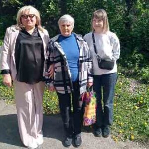 Троє жінок стоять поруч на асфальтованій доріжці на сонячному подвір’ї, оточеному густою зеленою рослинністю. Посередині — літня жінка в синій кофті та картатій куртці, тримає яскраву кольорову сумку.
