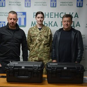 Троє людей стоять за столом із двома чорними захисними кейсами для обладнання на тлі брендованого банера Рівненської міської ради. Посередині – військовослужбовиця у камуфляжній формі, поруч із нею двоє чоловіків у темному одязі.