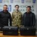 Троє людей стоять за столом із двома чорними захисними кейсами для обладнання на тлі брендованого банера Рівненської міської ради. Посередині – військовослужбовиця у камуфляжній формі, поруч із нею двоє чоловіків у темному одязі.