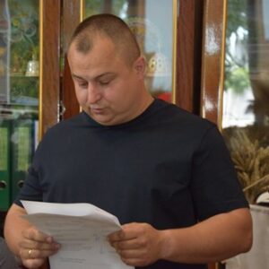 Чоловік у темній футболці стоїть у приміщенні та уважно читає документи, які тримає в руках. На задньому плані видно дерев’яну шафу зі скляними дверцятами, папки з документами та елементи декору.