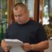 Чоловік у темній футболці стоїть у приміщенні та уважно читає документи, які тримає в руках. На задньому плані видно дерев’яну шафу зі скляними дверцятами, папки з документами та елементи декору.