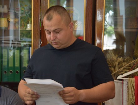 Чоловік у темній футболці стоїть у приміщенні та уважно читає документи, які тримає в руках. На задньому плані видно дерев’яну шафу зі скляними дверцятами, папки з документами та елементи декору.