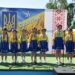 Група дітей у синьо-жовтих українських народних костюмах виступає на сцені просто неба.