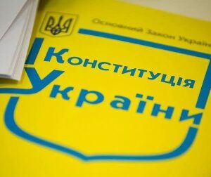 Жовто-блакитна обкладинка Конституції України лежить на столі поруч із паперовими документами. На обкладинці зображено Державний Герб України та великий напис «Конституція України»