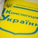 Жовто-блакитна обкладинка Конституції України лежить на столі поруч із паперовими документами. На обкладинці зображено Державний Герб України та великий напис «Конституція України»