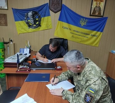 У службовому кабінеті за столом сидять двоє чоловіків — один у цивільному одязі, інший у військовій формі — та підписують документи про співпрацю.