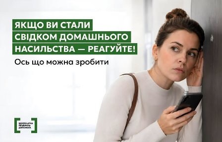 Жінка у світлому одязі стоїть біля стіни, тримаючи в руці смартфон і насторожено прислухаючись.