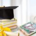 На столі розміщені стопка книг із академічною шапочкою, диплом із жовтою стрічкою та пачка українських гривень. Зображення символізує освіту, студентські досягнення, стипендіальну підтримку та фінансове заохочення здобувачів освіти.