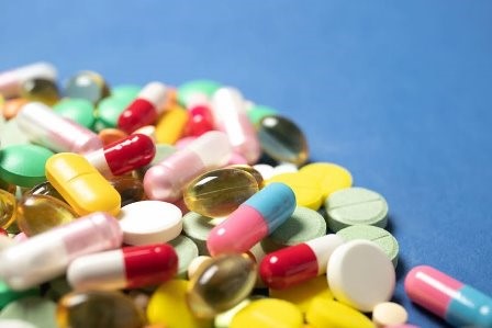 На синьому фоні розсипані різнокольорові лікарські засоби: капсули, таблетки та прозорі желатинові капсули овальної форми. Зображення символізує медикаменти, фармацевтичну продукцію та лікування.