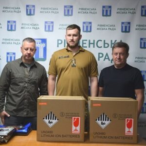 Троє чоловіків стоять у приміщенні Рівненської міської ради на тлі брендованого банера та прапорів України й міста Рівного.