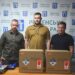 Троє чоловіків стоять у приміщенні Рівненської міської ради на тлі брендованого банера та прапорів України й міста Рівного.