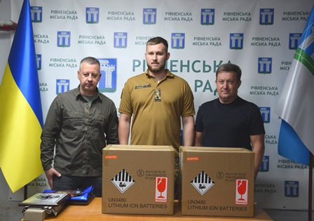 Троє чоловіків стоять у приміщенні Рівненської міської ради на тлі брендованого банера та прапорів України й міста Рівного.