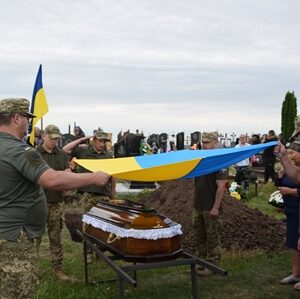 Військовослужбовці під час церемонії поховання тримають над труною загиблого Захисника України синьо-жовтий Державний Прапор.