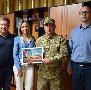 Четверо людей стоять в службовому кабінеті. У центрі — військовослужбовець у камуфляжній формі, який тримає в руках подяку в рамці.