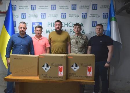 П'ятеро чоловіків стоять за столом у приміщенні Рівненської міської ради. На столі розміщено три великі коробки з обладнанням Starlink.