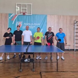У спортивній залі шестеро учасників турніру з настільного тенісу стоять за тенісним столом, тримаючи ракетки.