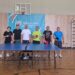 У спортивній залі шестеро учасників турніру з настільного тенісу стоять за тенісним столом, тримаючи ракетки.