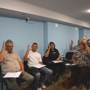 У залі засідань кілька учасників піднімають руки під час голосування. Люди сидять на стільцях із документами в руках.