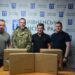 На фото четверо чоловіків стоять у приміщенні на тлі символіки Рівненської міської ради. На передньому плані розміщені дві великі картонні коробки з маркуванням, імовірно з допомогою для військових.