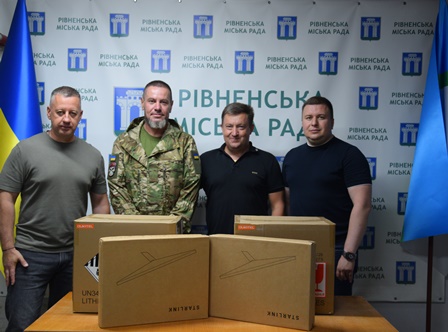 На фото четверо чоловіків стоять у приміщенні на тлі символіки Рівненської міської ради. На передньому плані розміщені дві великі картонні коробки з маркуванням, імовірно з допомогою для військових.
