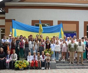 Групове фото учасників святкового заходу перед будівлею. Люди різного віку стоять і сидять разом на тлі великого синьо-жовтого Державного Прапора України; частина учасників у вишиванках, поруч — букети соняшників та квіти.