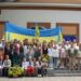 Групове фото учасників святкового заходу перед будівлею. Люди різного віку стоять і сидять разом на тлі великого синьо-жовтого Державного Прапора України; частина учасників у вишиванках, поруч — букети соняшників та квіти.