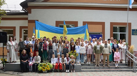 Групове фото учасників святкового заходу перед будівлею. Люди різного віку стоять і сидять разом на тлі великого синьо-жовтого Державного Прапора України; частина учасників у вишиванках, поруч — букети соняшників та квіти.