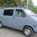 Сірий мікроавтобус із багажником на даху припаркований на вулиці біля будівель та дерев.