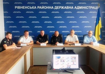 П’ятеро чоловіків беруть участь в онлайн-зустрічі в приміщенні Рівненської районної державної адміністрації. На столі перед ними — таблички з іменами та комп’ютерний монітор із відеоконференцією.