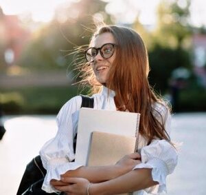 Дівчина в окулярах із книгами та зошитами в руках стоїть на вулиці.