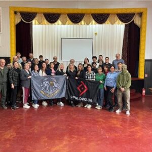 Групове фото учасників навчання у приміщенні Будинку культури. Люди стоять разом перед екраном, тримаючи прапори та символіку організацій.