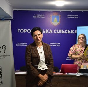 Дві жінки під час заходу в приміщенні Городоцької сільської ради; на фоні — банер громади та презентація програми «Ти як? — Маршрути підтримки кожного в громаді».