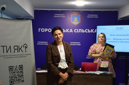 Дві жінки під час заходу в приміщенні Городоцької сільської ради; на фоні — банер громади та презентація програми «Ти як? — Маршрути підтримки кожного в громаді».