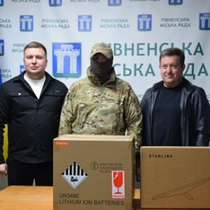 На фото троє чоловіків стоять біля столу на тлі стенда Рівненської міської ради та українського прапора.