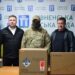 На фото троє чоловіків стоять біля столу на тлі стенда Рівненської міської ради та українського прапора.