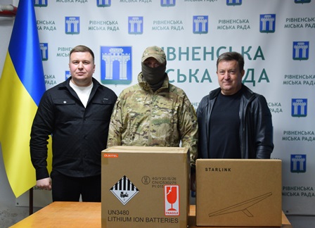 На фото троє чоловіків стоять біля столу на тлі стенда Рівненської міської ради та українського прапора.