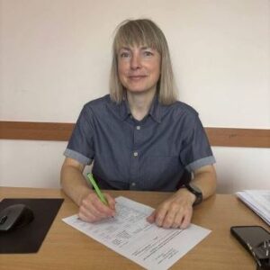 Жінка сидить за робочим столом в офісі та заповнює документи. На столі лежать папери, ручки та інші канцелярські речі.