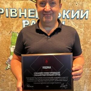 Чоловік стоїть у приміщенні та тримає в руках пам’ятну подяку в рамці. На задньому плані — карта та напис українською мовою.
