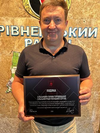 Чоловік стоїть у приміщенні та тримає в руках пам’ятну подяку в рамці. На задньому плані — карта та напис українською мовою.