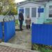 На фото двоє людей стоять біля входу на територію Городоцької амбулаторії. Вхід облаштований синіми металевими воротами, поруч розташована будівля закладу з інформаційною табличкою.