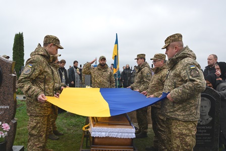 Військовослужбовці тримають Державний Прапор України над труною під час церемонії прощання із загиблим Захисником України на кладовищі.