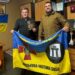 Двоє чоловіків тримають прапор військової частини 3055 Національної гвардії України. Чоловік ліворуч тримає пам’ятну відзнаку.